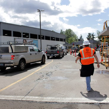 A worker wearing a reflective vest and hardhat walks near a line of trucks waiting to enter a large industrial building