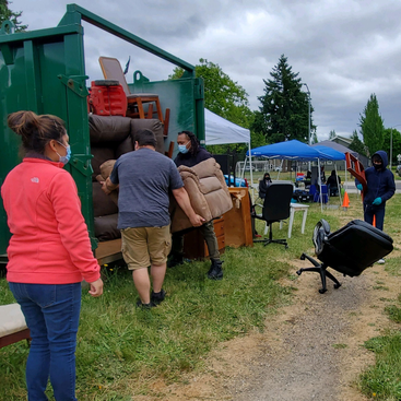 A group of people load pieces of furniture into a large green metal storage unit on a grassy field along a street in Portland