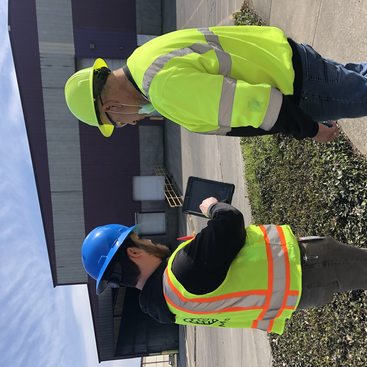 Two men wearing hard hats and reflective safety vests review an electronic tablet during an enforcement inspection to a solid waste transfer station in greater Portland