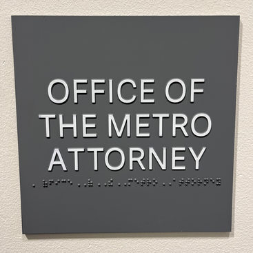 A dark gray wayfinding sign with white letters and Braille text that reads Office of the Metro Attorney