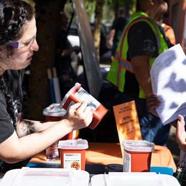 One woman shows a quart-sized sharps container to another woman while at an event. A man in a safety vest stands in the background. 