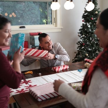 Three people around a dining room table investigating wrapped presents with a Christmas tree in the background.