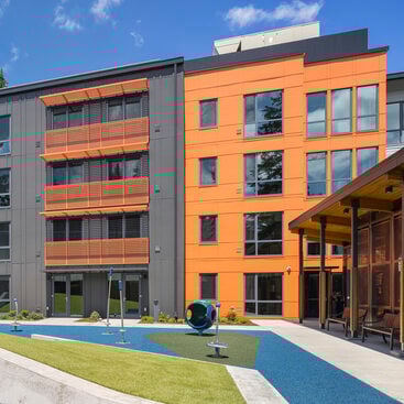 A panoramic view of the Wynne Watts apartment complex on a sunny day from the perspective of the interior courtyard, with a children's play area in the foreground. 