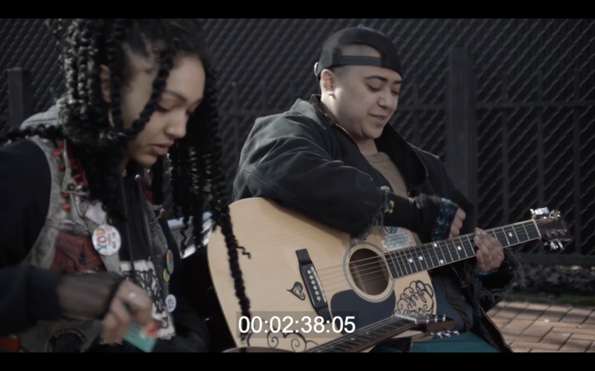 screenshot from film showing two youths sitting down with one playing guitar