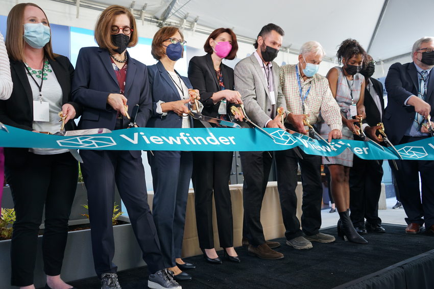 Nine people standing on a stage and cutting a large ribbon with oversized scissors