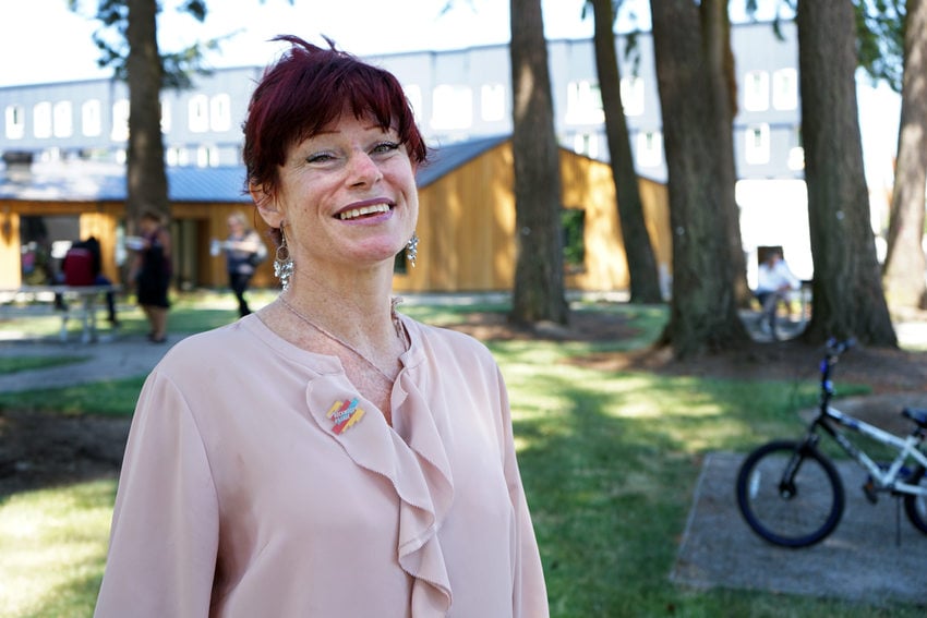 White woman in a pink ruffled shirt with red hair and bangs, smiling, in a park with tall trees and multifamily housing in the background