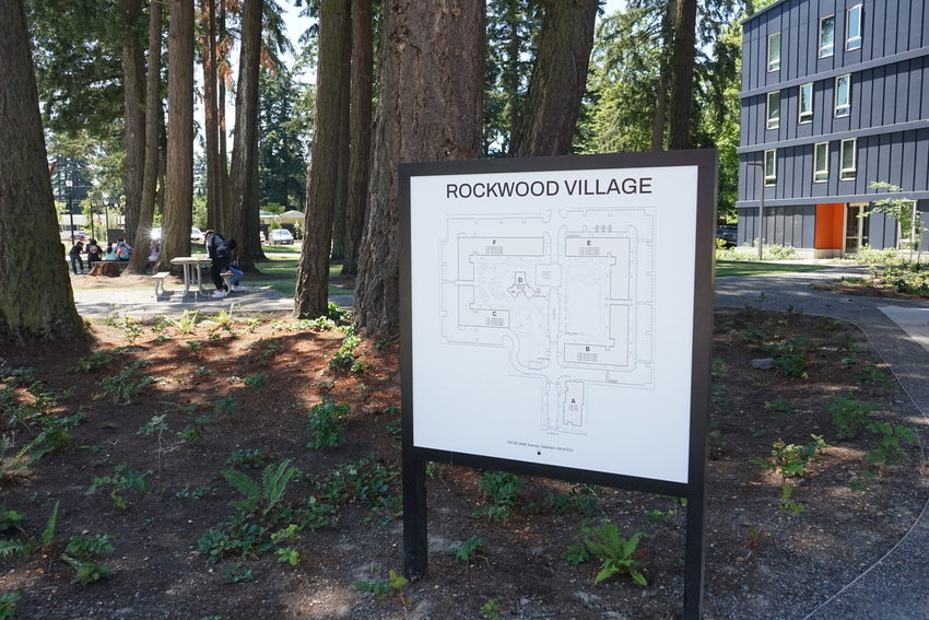 Sign with Rockwood Village residential complex site plan in foreground. Tall trees and picnic tables in background, with a dark blue multifamily building in background on the right