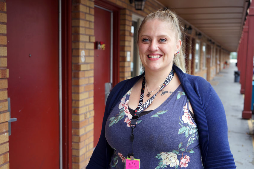 Blonde white woman in blue cardigan standing in exterior motel corridor smiling