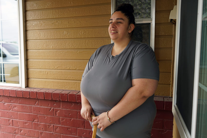 Woman standing in front of building exterior inner corner, wearing grey shirt, smiling and holding a cane