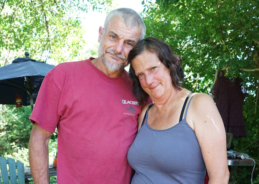 Portrait of a couple in an outdoor setting: Man with red shirt and short grey hair and beard with a woman in a blue tank top with chin length brown hair.