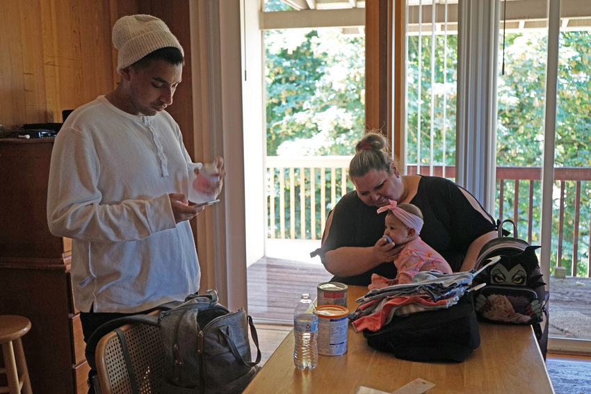 Young man in white shirt and beanie stands looking at baby bottle, while woman in black blouse gives a pacifier to a baby seated on the table.