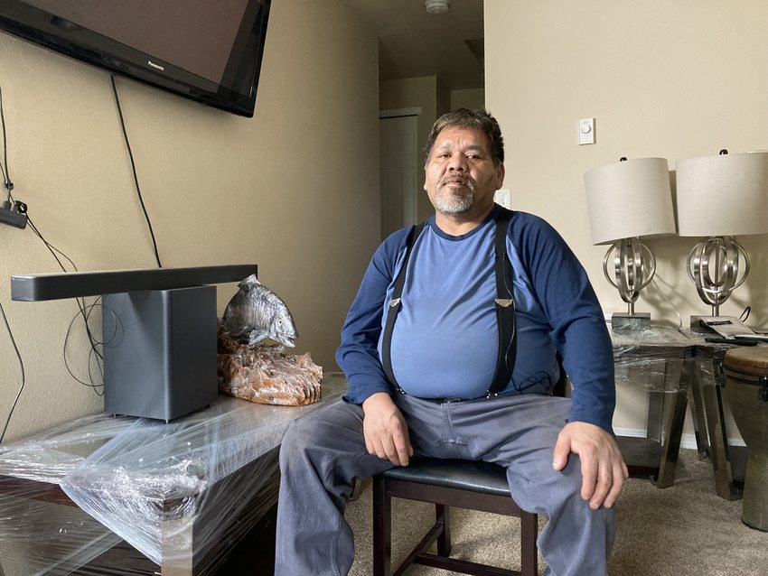 Man in a blue shirt with suspenders sitting in an apartment with various furnishings in the background, including a ceramic salmon