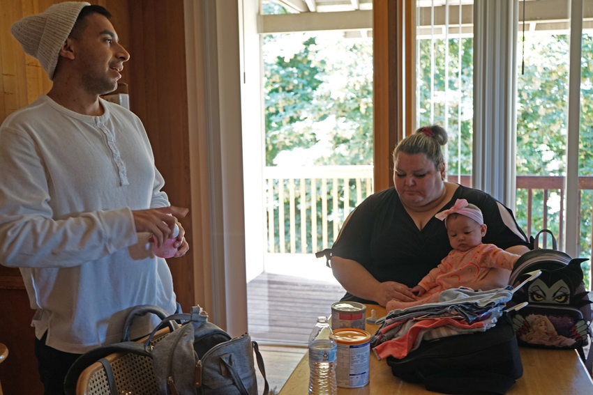 Man in a white shirt and beanie hat standing and smiling; woman in a black dress sitting at a table holding a baby in a pink jumper