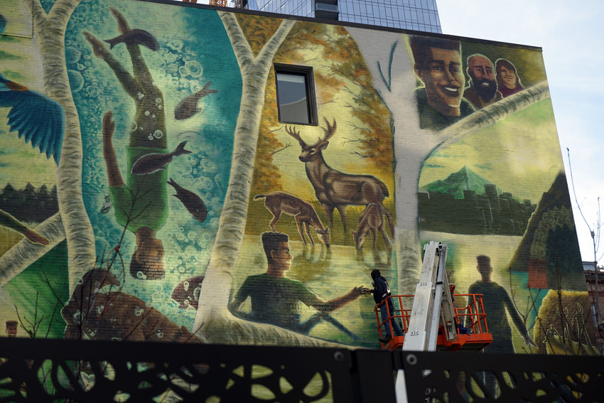 Mural of several scenes with a Black man in natural settings, encountering fish, a feer and a mountain landscape. The artist is seen working on it in the lower right corner.