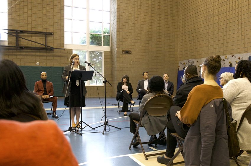 Metro Councilor Christine Lewis stands at a lectern and speaks before a crowd in a large gymnasium with a bright window behind her.