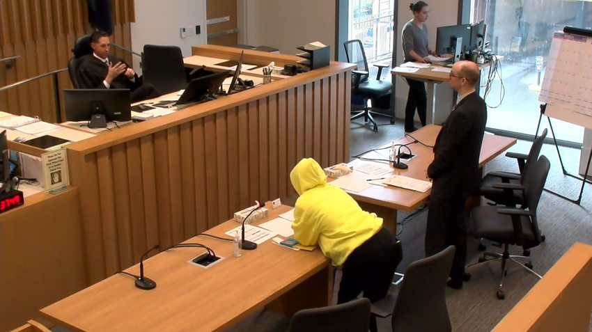 Court room with a judge addressing a person in a suit and another person in a bright yellow hooded sweatshirt