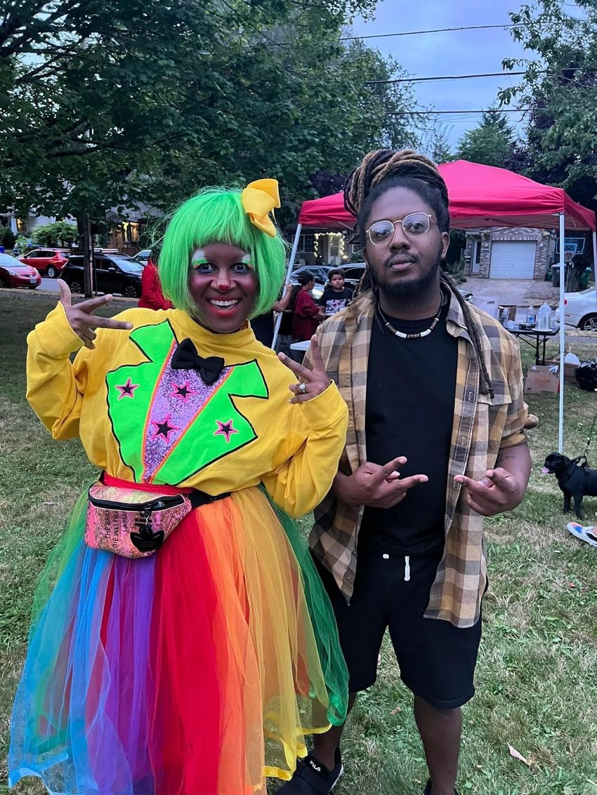 On the left side, Nikki Brown Clown flashes two peace signs in her colorful costume as she poses with event organizer Donovan Scribes on the right who flashes two peace signs