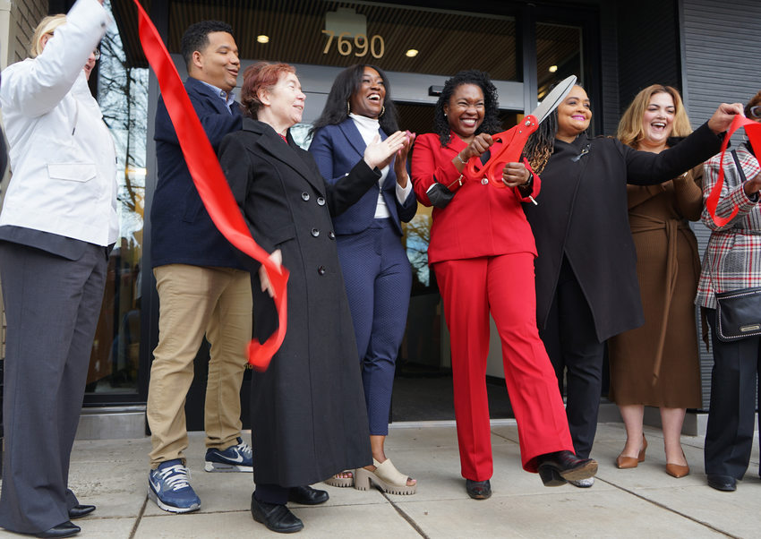 Eight people just after cutting a large red ribbon in front of an apartment building. A woman in the center in a red suit holds oversized red scissors