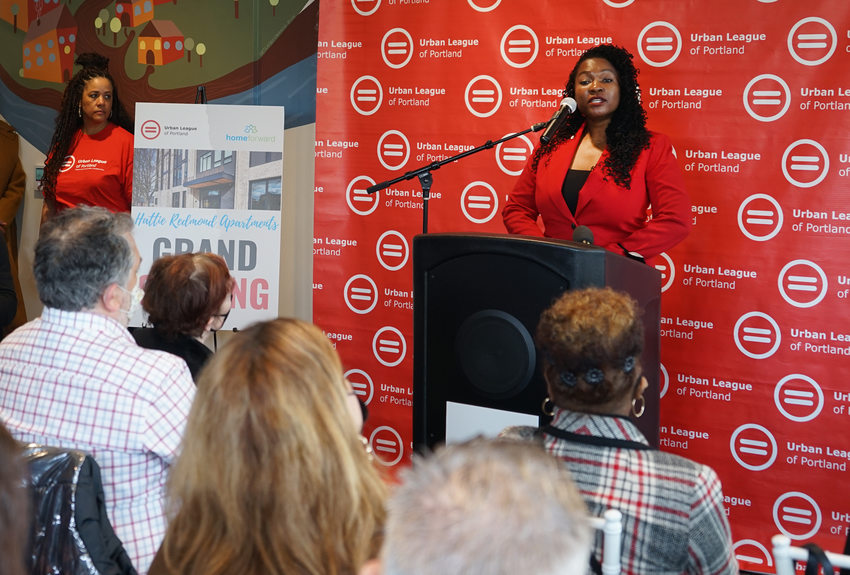 Woman in a red blazer speaking at a podium to a small crowd. The backdrop reads "Urban League of Portland"