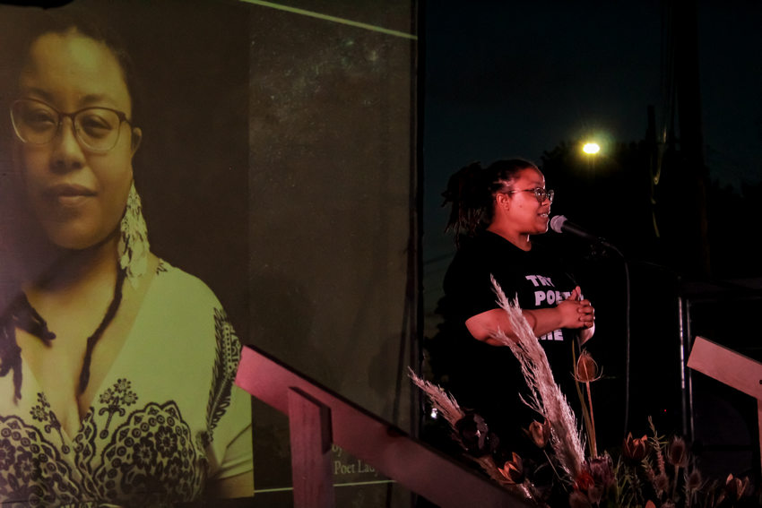 A Black woman performs poetry on stage at night time with a large photo of her on the projector in the background