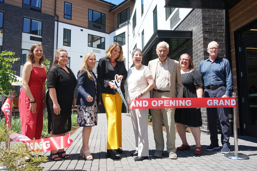 Eight people posing for a photograph just after cutting a red "grand opening" ribbon with giant scissors.