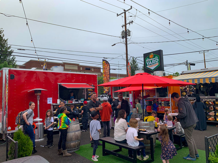 People of all ages gather at The Yard food cart pod during an overcast, rainy day. Some stand while others sit at tables underneath red umbrellas.