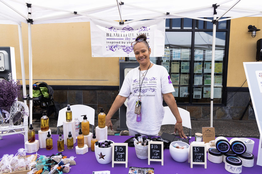 A light brown skinned woman stands in her booth behind a table covered in a purple tablecloth and jars of white lotions, golden body oils, lip glosses and other handmade vegan cosmetics.