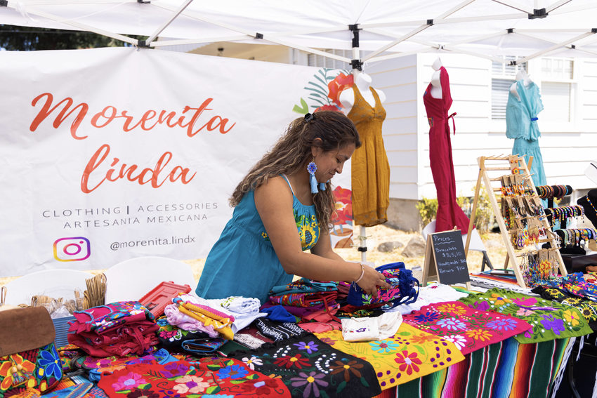 A young light brown skinned woman places blue embroidered bags on a table full of other embroidered items.