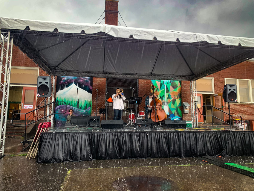 A medium skinned man plays the trombone and a light-skinned man plays the cello on stage at Vestal Social Justice Night as rain pores down on the blacktop.