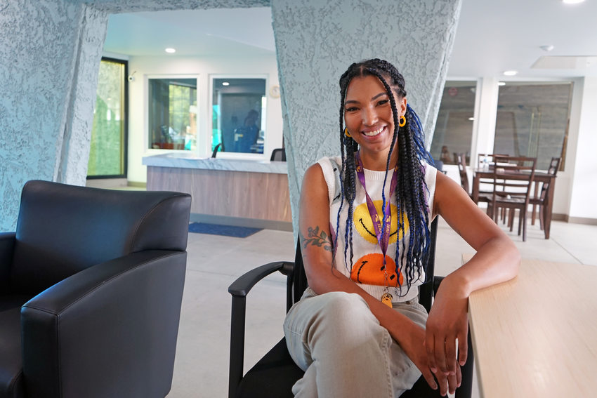 Young woman with long, multicolored braids smiling and sitting by a table in an apartment building lobby.
