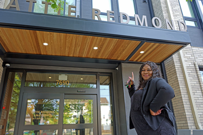 Woman dressed in all black standing in front of an apartment building smiling and pointing up at a sign that reads "Hattie Redmond" with the 'H' just out of frame.