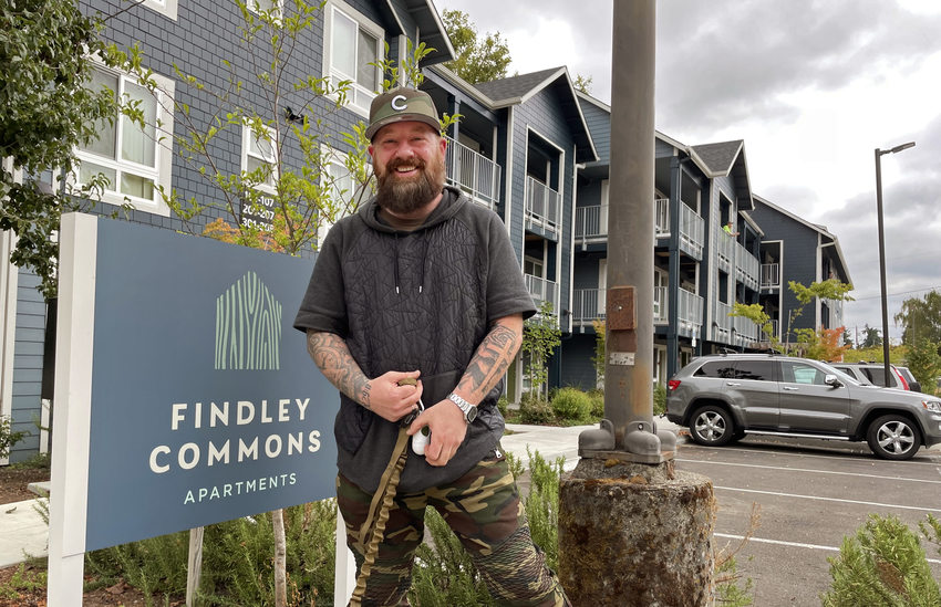 Man wearing a sweatshort and camo pants, holding a dog leash and smiling by a sign that reads "Findley Commons Apartments" in front of an apartment building.