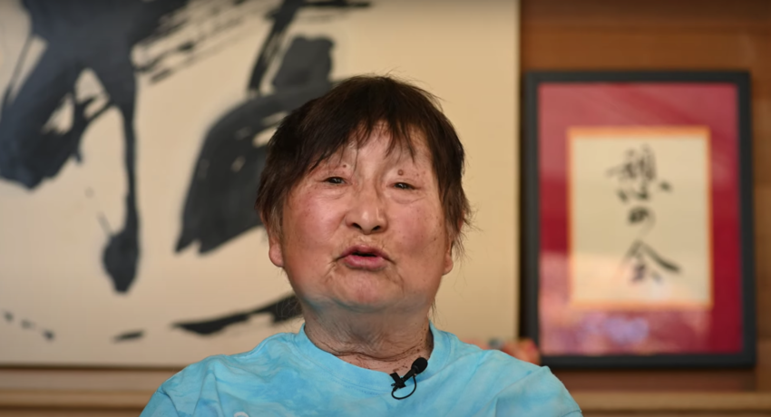 An elderly Japanese woman with short brown hair wearing an aquamarine t-shirt sits in front of abstract Japanese calligraphy art