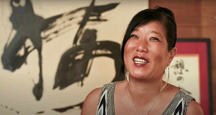 A young Japanese-American woman with black hair in a bun, wearing a black and white stripped tank top, sits in front of abstract Japanese calligraphy art. 