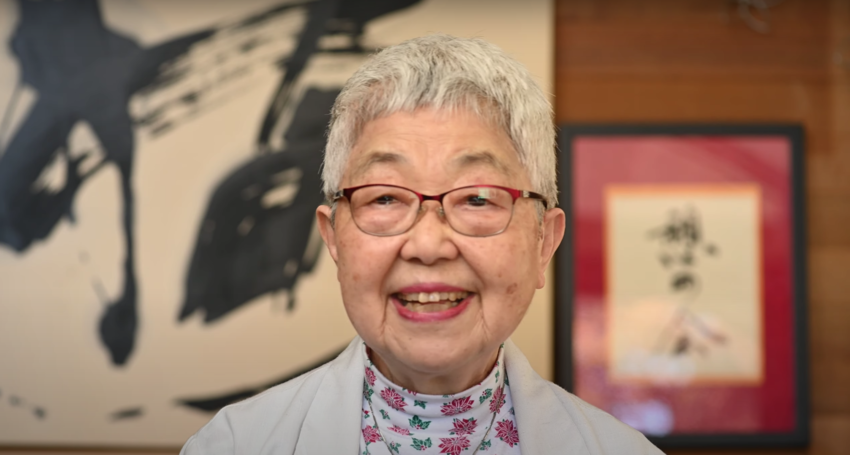 An elderly Japanese woman with short grey hair wearing glasses and a white floral shirt an sits in front of abstract Japanese calligraphy art.
