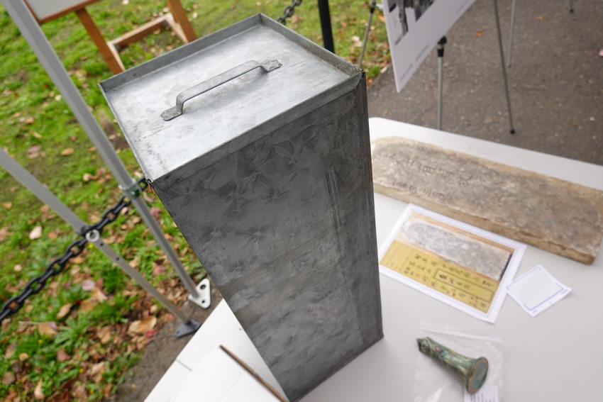 A tall skinny rectangular metal box stands on a table next to a gravestone inscribed in Chinese writing and a green porcelain object