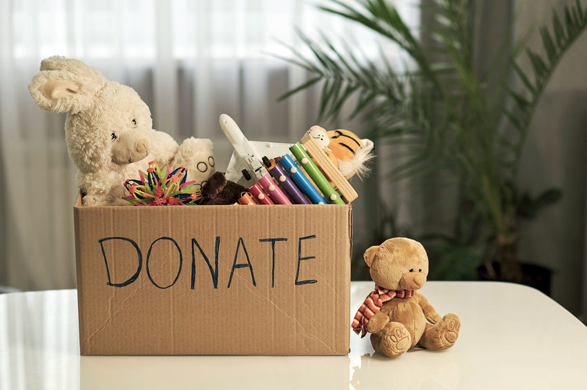 A cardboard box full of toys is on a table next to a teddy bear, the box reads donate in large letters