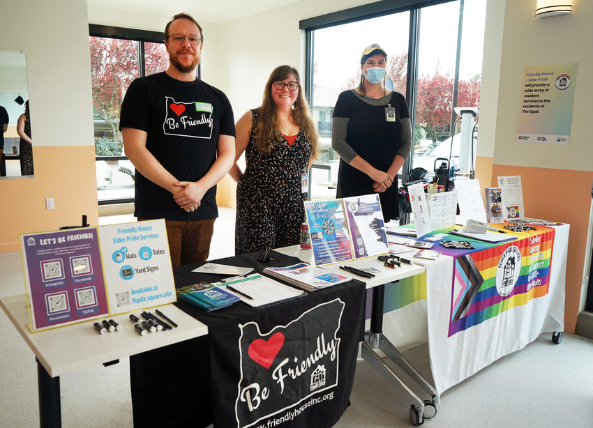 Three people standing behind a table with informational materials and a banner reading "Be Friendly."