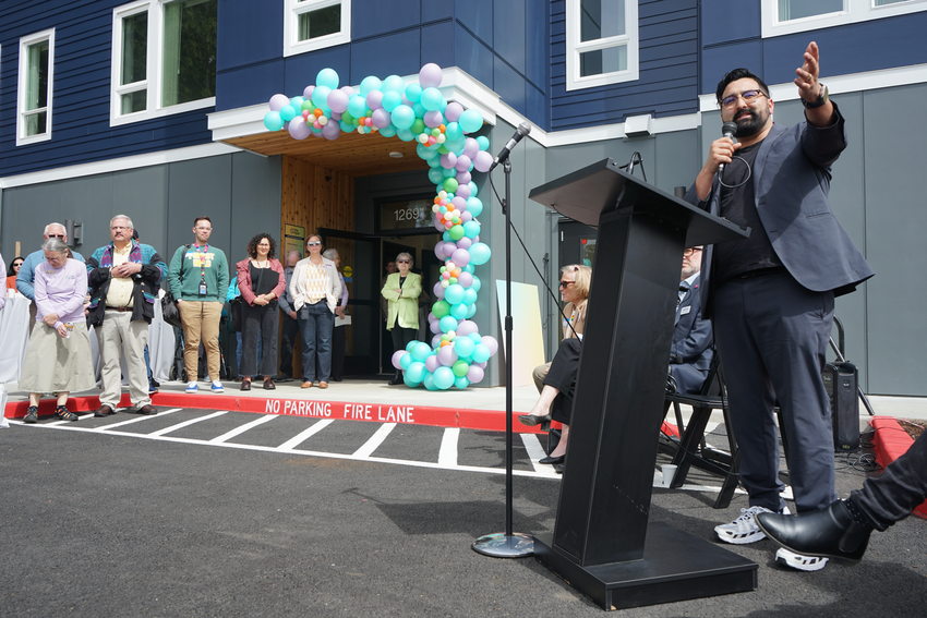 Man in a suit speaking at a podium with an audience to his left and balloons decorating the doorway of an apartment complex they are standing in front of.