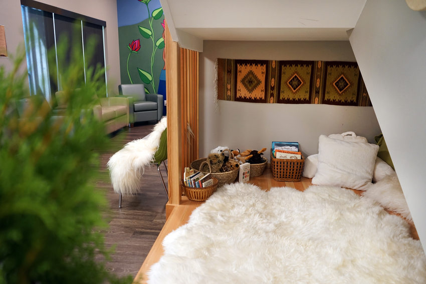 Cozy reading nook under a slanted ceiling with a plush white fur rug and a low bed surrounded by woven baskets of books and stuffed animals. A room with a sofa and a mural of large flowers is visible through an opening on the left. 