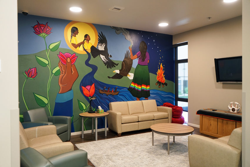 A community room with a large mural depicting various scenes with revelvance to Native peoples: an elder and child surrounded by flowers, an eagle in flight, and a group in a canoe on a river. 