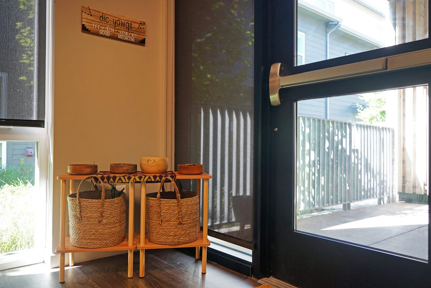 The corner of a room with a wood rack containing several baskets. A sign above reads "good medicine" in English and Chinook. To right there's the door to a sunny patio. . 