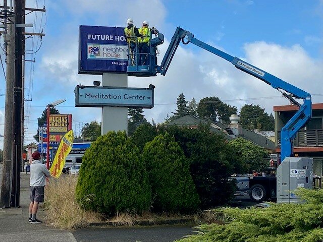 A sign for Neighborhood House is installed by workers in a forklife