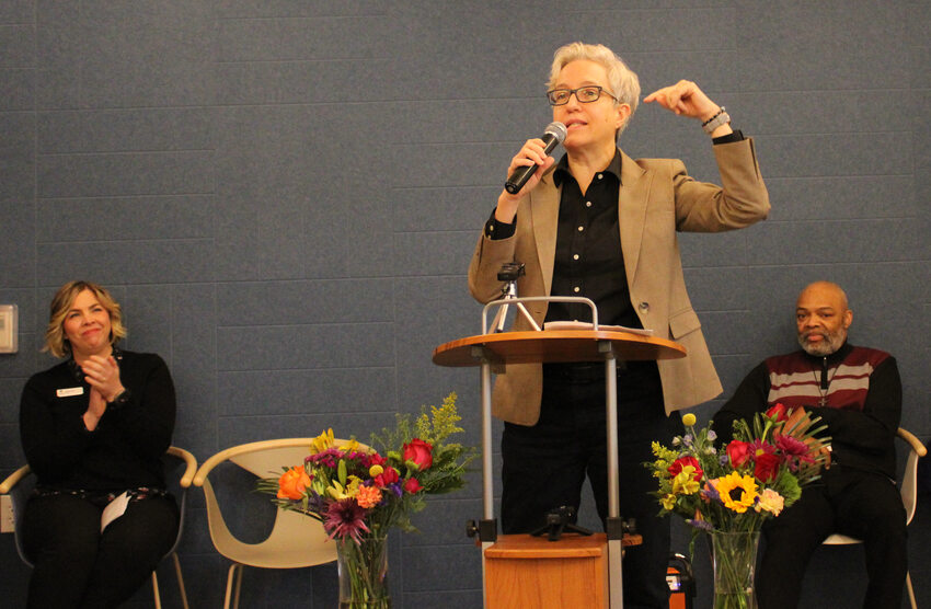 Woman with short, light colored hair and wearing a black shirt with a brown blazer speaks emphatically at a podium. 