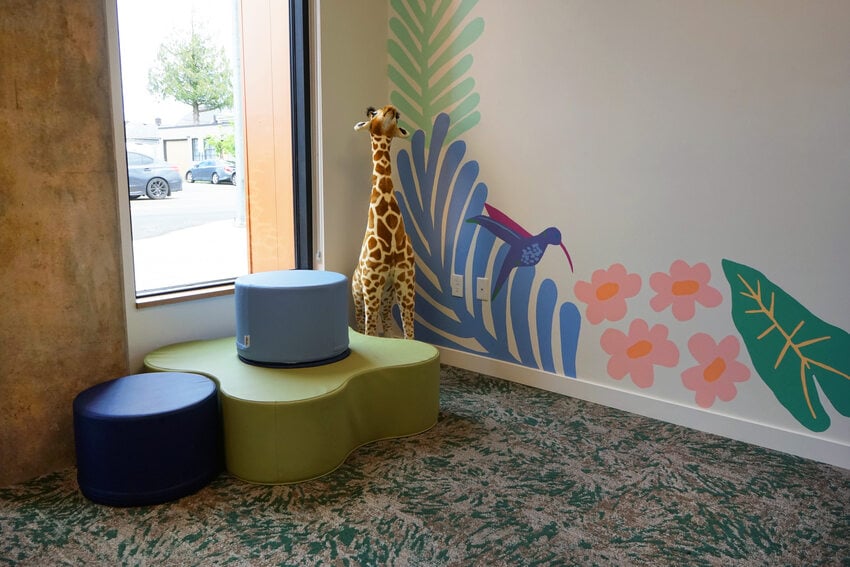 Corner of a room with colorful seating objects and a stuffed giraffe beside a colorful mural 