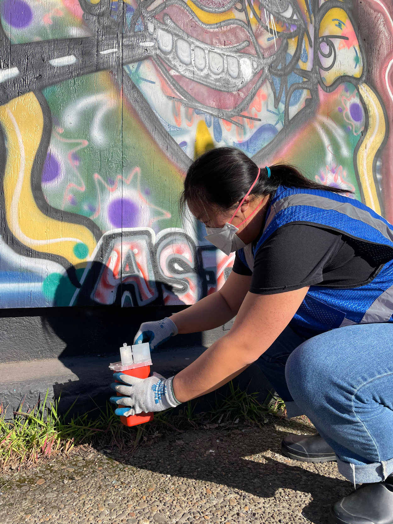 An image of volunteer kneeling to pick up sharps in front of a graffitied wall.