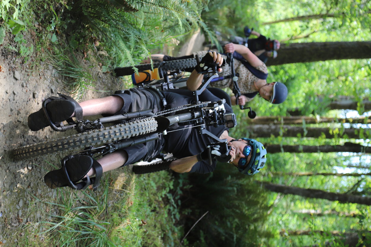 an adaptive mountain biker rides down a dirt trail with a person on a standard bike behind him