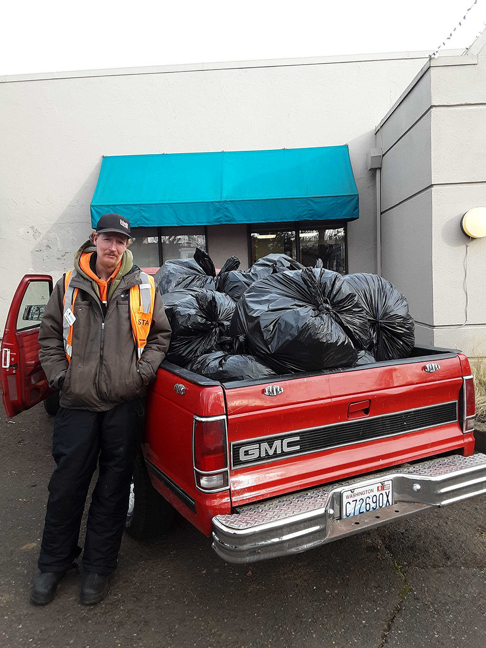 A man leans against a red pickup truck. The bed it full of filled garbage bags.