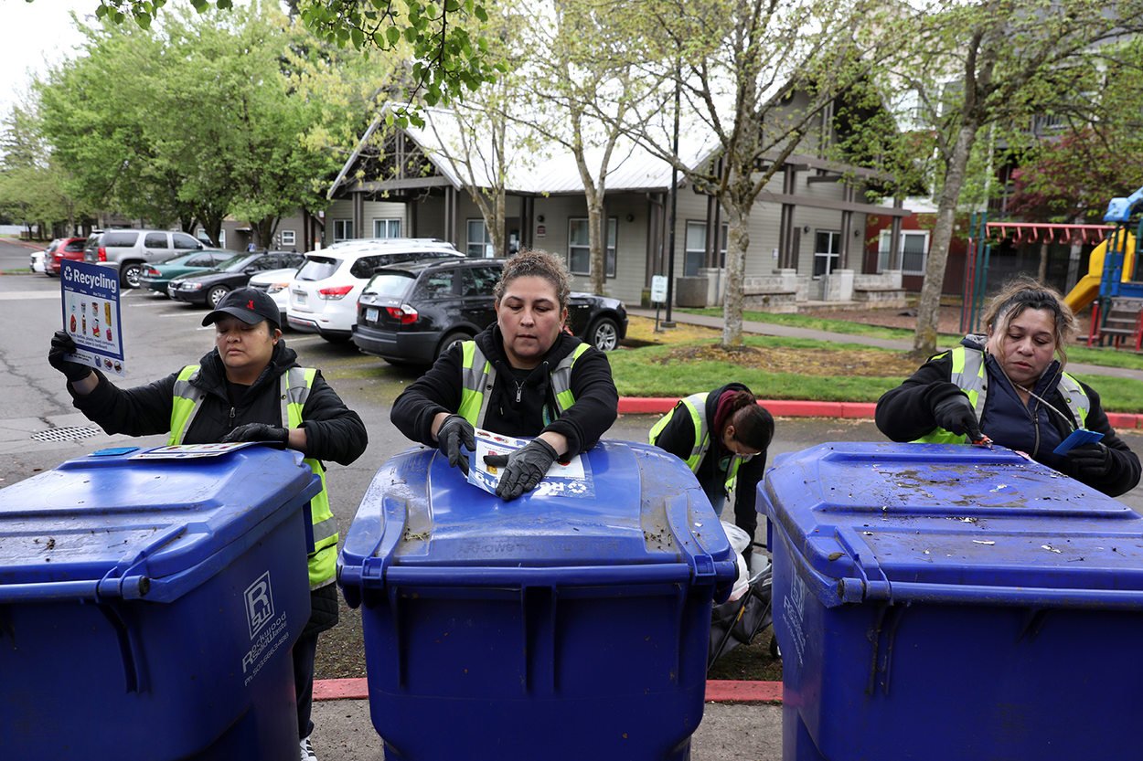 Three woman stand infront of three recycling bins, all of them applying new decals to the outside of the bin