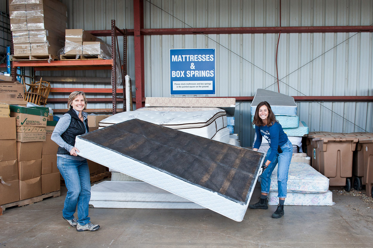 Two women carry a mattress to a pile of mattresses for recycling.
